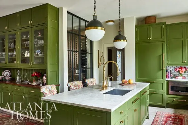 Green kitchen with marble island, brass hardware, glass-front cabinets, and a wine cellar visible through a glass wall.