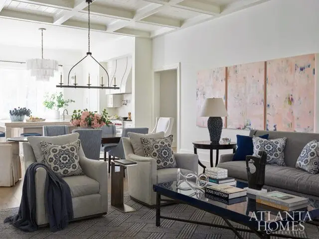 Living room with gray sofa and armchairs, blue and white patterned pillows, coffered ceiling, and abstract triptych art above a lamp on a side table.