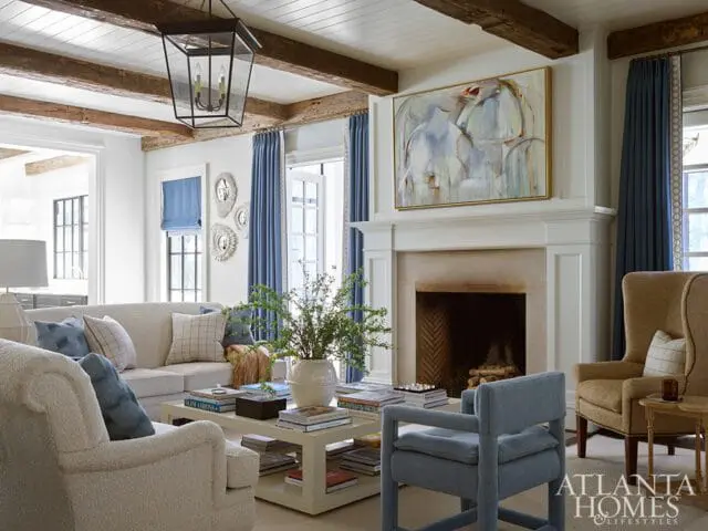 Living room with white walls, exposed wood beams, a stone fireplace with abstract art above, and upholstered furniture in neutral tones and blue accents.