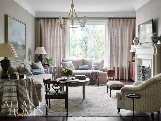 Living room with neutral-toned sofas, patterned curtains, a round coffee table, and a fireplace with decorative urns.