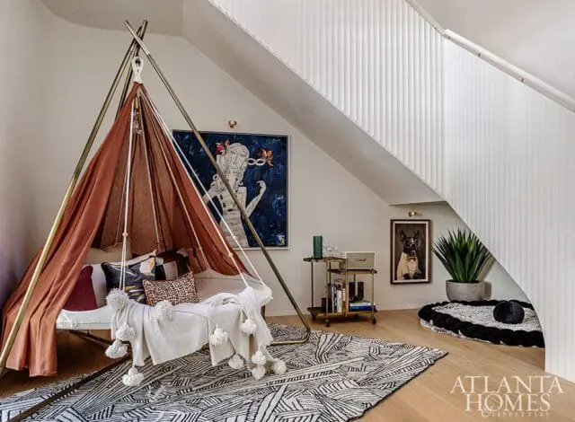 Cozy reading nook with a copper-colored tent structure, daybed with pillows, and a black and white geometric rug. Artwork and a bar cart are visible nearby.