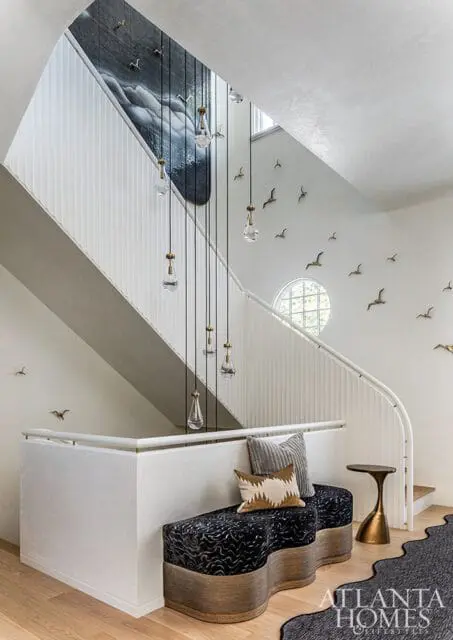 Foyer with a curved staircase, white paneled walls, and a large abstract mural. A series of glass pendant lights hang down the stairwell. Decorative bird sculptures are mounted on the walls.