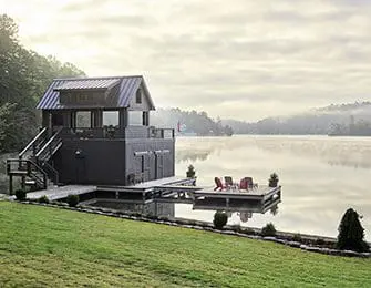 Dark gray boathouse with metal roof on a lake, featuring a deck with chairs.