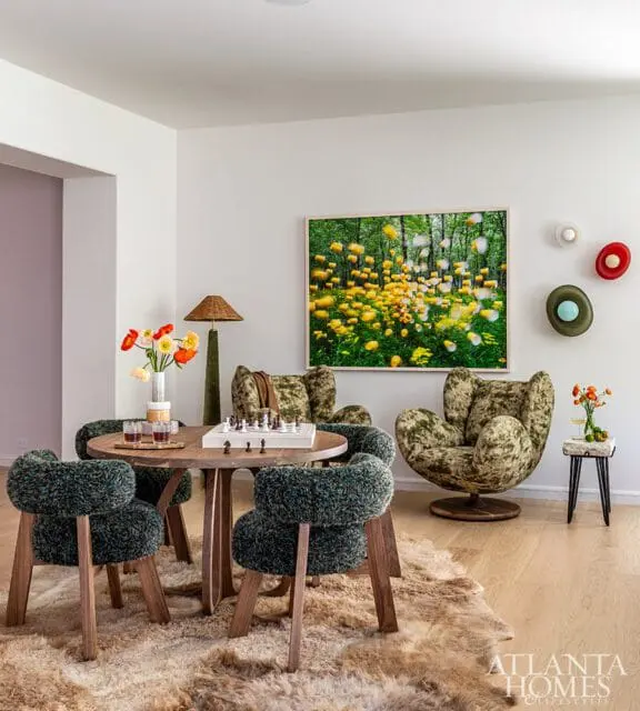 Game room with round wood table, four textured green chairs, two plush armchairs, and a large abstract painting of yellow flowers in a forest.