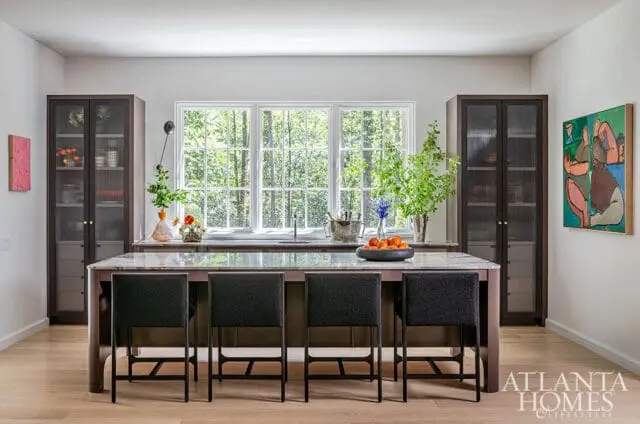 Modern kitchen with a large island, dark cabinetry, marble countertops, and a wall of windows overlooking trees.