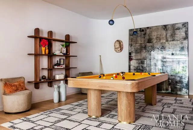 Game room with a light wood pool table with yellow felt, a sculptural wood shelf, a textured armchair, and a large distressed mirror wall.