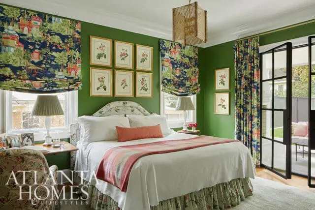 Green bedroom with a white bed, floral patterned curtains and roman shades, and framed botanical prints on the wall.