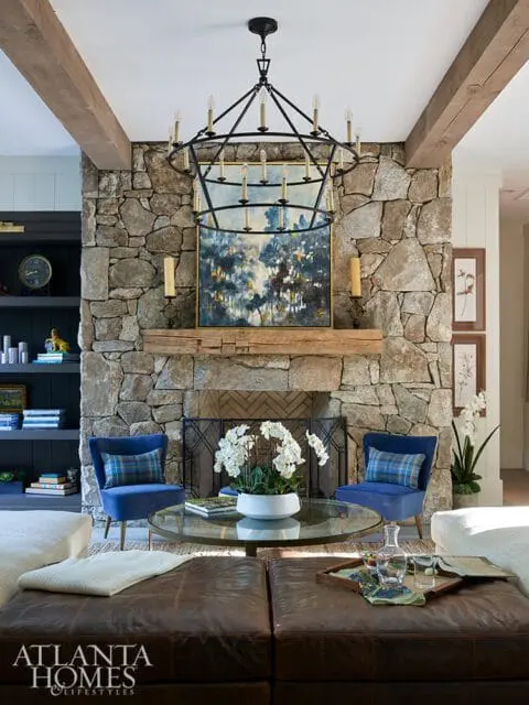 Living room with stone fireplace, wooden mantel, large circular chandelier, blue chairs, glass coffee table, and leather ottomans.