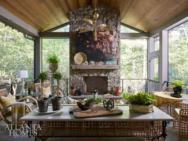 Screened porch with stone fireplace, wood-paneled ceiling, wicker furniture, and a marble-top table. Lush greenery is visible through the screens.