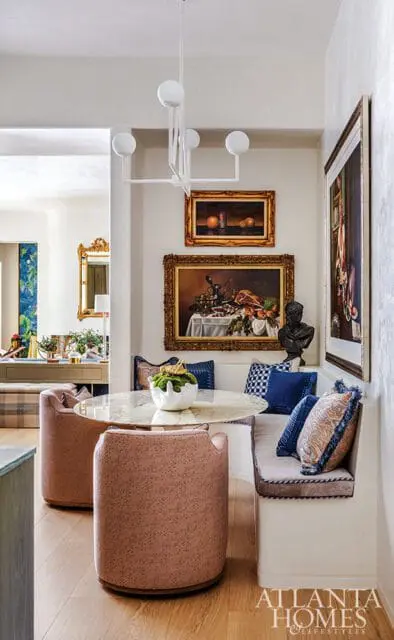 Dining nook with a marble-topped table, pink upholstered chairs, built-in banquette seating with blue and patterned pillows, and still life paintings on the wall.