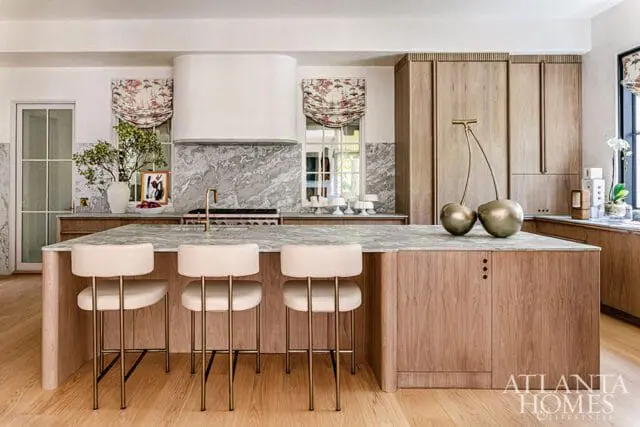 Modern kitchen with a large marble island, light wood cabinetry, and cream upholstered bar stools. A white range hood is centered above a marble backsplash.