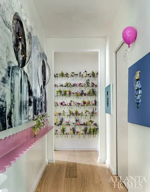 Hallway with a large black and white portrait on the left, a pink scalloped shelf with a vase of flowers, and a wall display of numerous small flower arrangements.