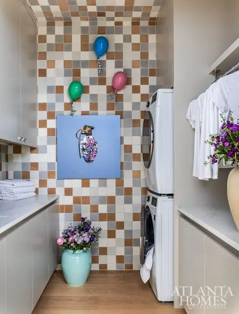 Modern laundry room with stacked white washer and dryer, light gray cabinetry, marble countertops, and a tiled backsplash featuring a colorful grenade artwork and balloons.