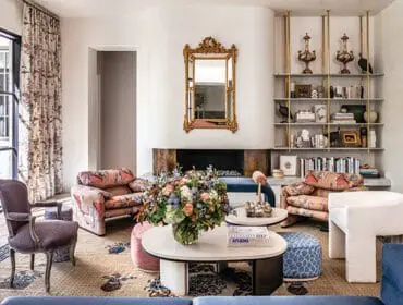 Living room with patterned armchairs, a floral rug, a gold-framed mirror above a fireplace, and built-in shelving filled with books and decorative objects.