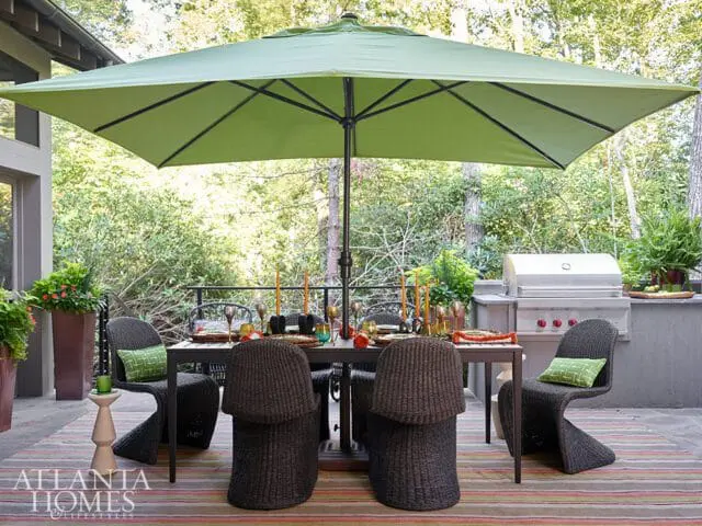 Outdoor dining area with a table set for four, wicker chairs, a green umbrella, and a built-in grill on a patio with a striped rug.