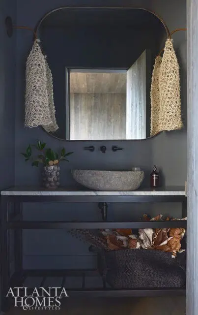 Dark blue bathroom with a stone vessel sink, marble countertop, and metal vanity. Woven baskets hang from a curved rod.