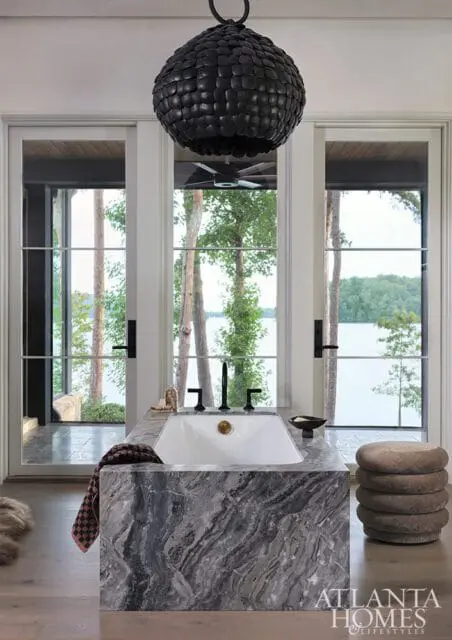 Bathroom with a large marble vanity featuring a white sink and black faucet, overlooking a lake through glass doors. A black pendant light hangs above.