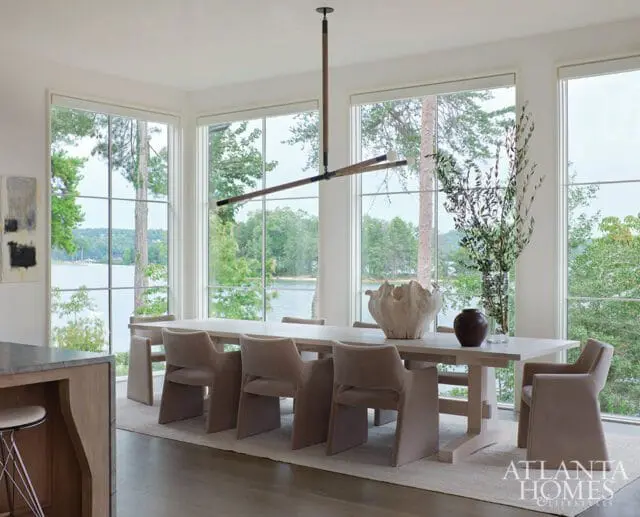 Dining room with a long wooden table, taupe upholstered chairs, and large windows overlooking a lake and trees.
