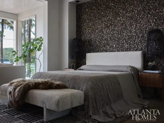 Bedroom with a textured dark wallpaper accent wall, a white upholstered headboard, and a bed dressed in gray linens. A fur throw drapes over a bench at the foot of the bed.