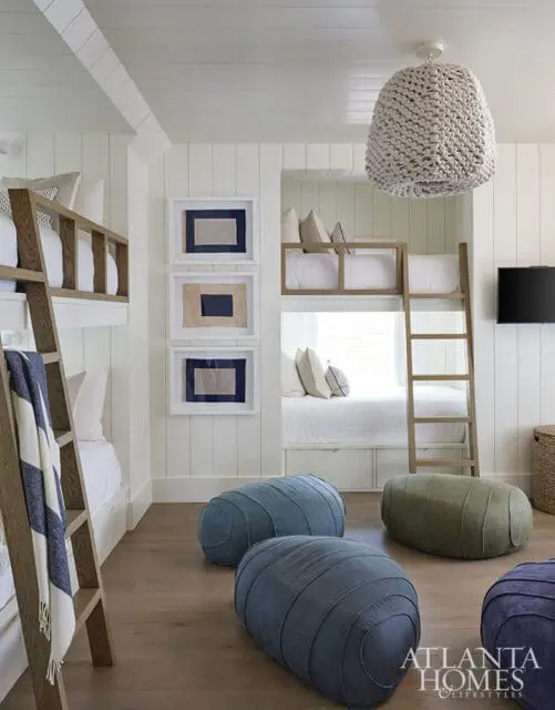 Bunk room with white shiplap walls, wooden ladders, framed nautical art, and plush floor cushions in blue, green, and purple.