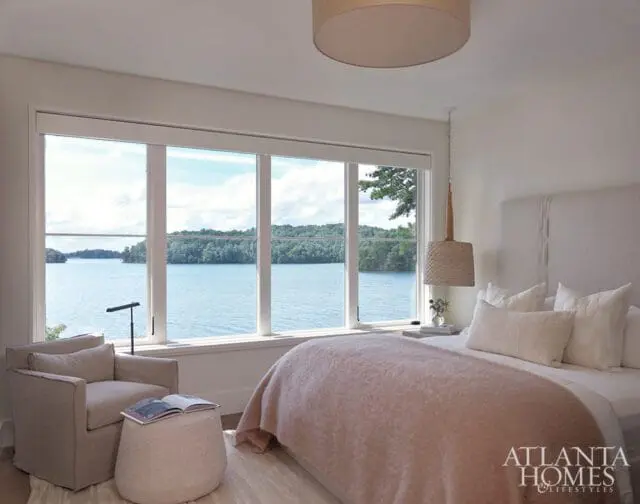 Bedroom with large windows overlooking a lake, featuring a neutral-toned armchair, ottoman, bed with blush pink throw, and woven pendant light.