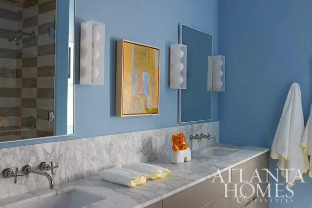 Blue bathroom with marble countertop, double sinks, modern chrome faucets, abstract art, and wall-mounted sconces.