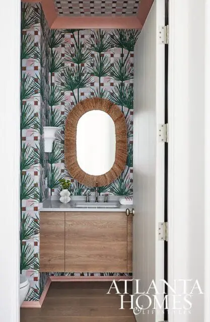Powder room with palm leaf wallpaper, a wooden vanity, oval mirror with woven frame, and a tiled ceiling.