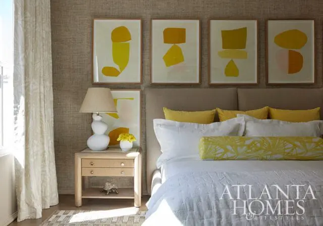 Bedroom with textured wallpaper, framed abstract art in yellow and orange, a white lamp on a light wood nightstand, and a bed with white linens and yellow accent pillows.