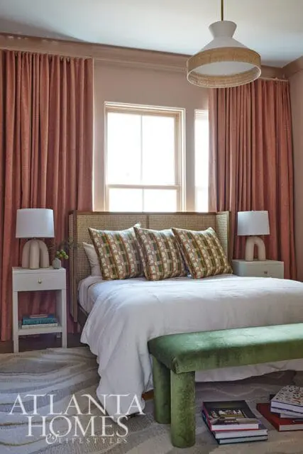 Bedroom with a cane headboard, white bedding, patterned pillows, coral curtains, and a green velvet bench at the foot of the bed.