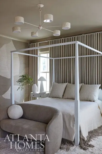 Bedroom with a white canopy bed, textured pillows, a curved sofa, and a modern chandelier. Neutral tones dominate the space.