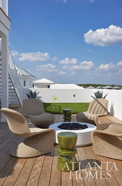 Outdoor living area with wicker chairs around a white fire pit, green ceramic stools, and a putting green in the background.