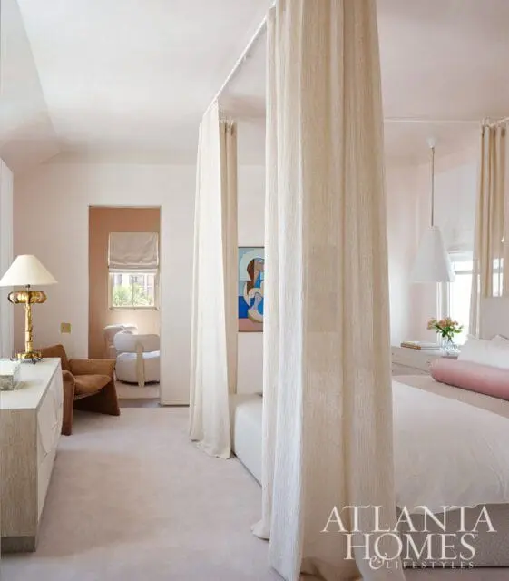 Bedroom with a four-poster bed draped in sheer curtains, a light wood dresser with a brass lamp, and a separate seating area visible through an opening.