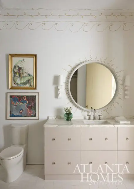 Bathroom with a light pink vanity, marble countertop, round mirror with sunburst detail, and two framed abstract paintings above a toilet.
