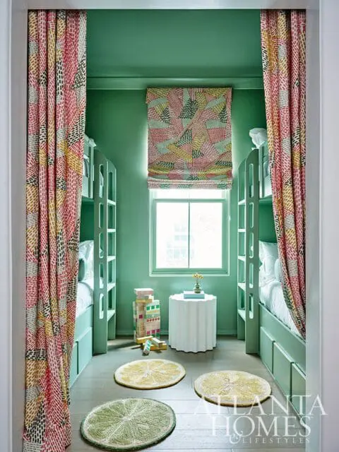 Bunk room with mint green walls and bunk beds, featuring colorful patterned curtains, roman shade, citrus-shaped rugs, and a fluted side table.