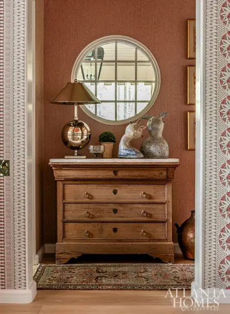 Mudroom with a wooden dresser topped with a marble slab, a globe lamp, decorative animal sculptures, and a round window with a grid pattern. The walls are covered in textured rust-colored wallpaper and patterned wallpaper borders.