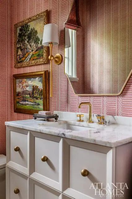Powder room with red patterned wallpaper, marble countertop, white vanity with brass hardware, and two framed landscape paintings.