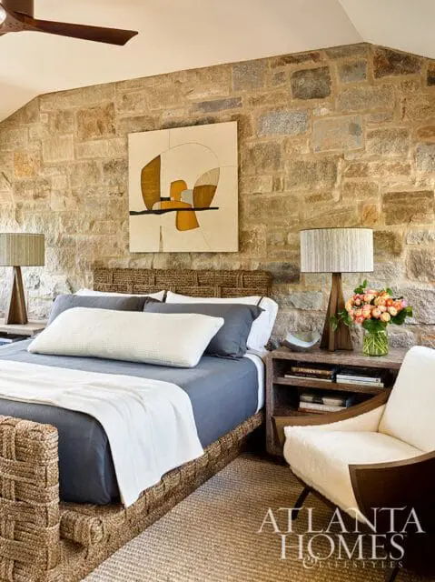 Bedroom with stone accent wall, woven bed frame, blue bedding, and abstract art above the headboard. A white armchair sits next to a nightstand with a vase of flowers.