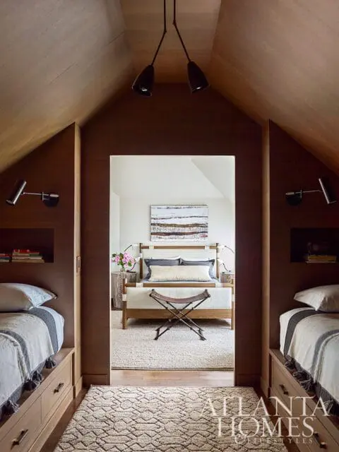 Bedroom with wood-paneled walls and vaulted ceiling, featuring two twin beds with built-in drawers and a doorway opening to a master bedroom with a king bed and abstract art.