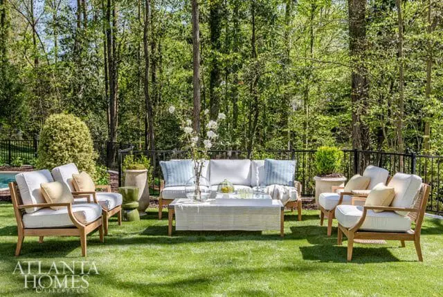 Teak outdoor furniture with white cushions and blue striped pillows arranged on a green lawn, backed by a dense forest and black metal fence.