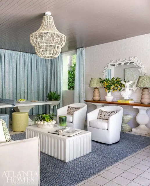 Living room with white swivel chairs, fluted white coffee table, blue rug, and light blue patterned curtains. A large woven chandelier hangs from the gray wood-paneled ceiling.