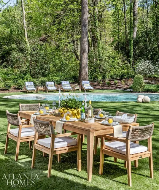 Teak dining table and chairs set on a green lawn with a swimming pool and lounge chairs in the background, surrounded by trees.
