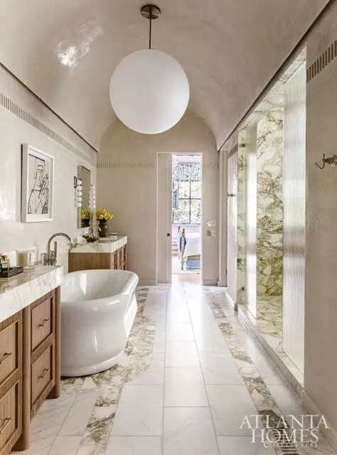 Primary bathroom with a freestanding tub, marble countertops, wood vanities, and a marble-tiled shower. A large spherical pendant light hangs from the vaulted ceiling.