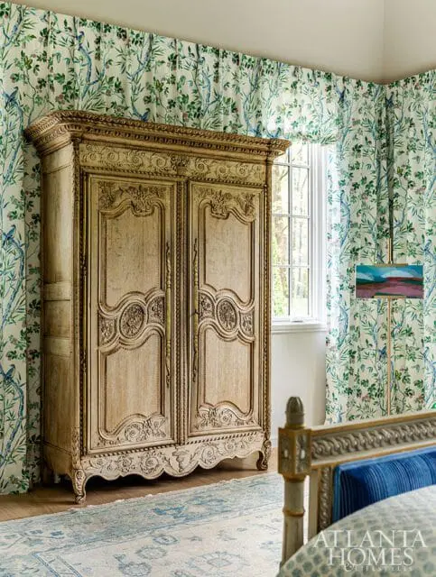 Primary bedroom with a vintage carved armoire, floral wallpaper, and a blue and white rug. A painting on an easel stands near the window.