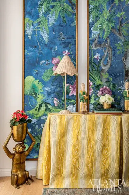 Primary bedroom with a gold monkey statue holding a planter, a table draped in a yellow and cream striped cloth, and two framed botanical murals featuring blue backgrounds, lush greenery, and butterflies.
