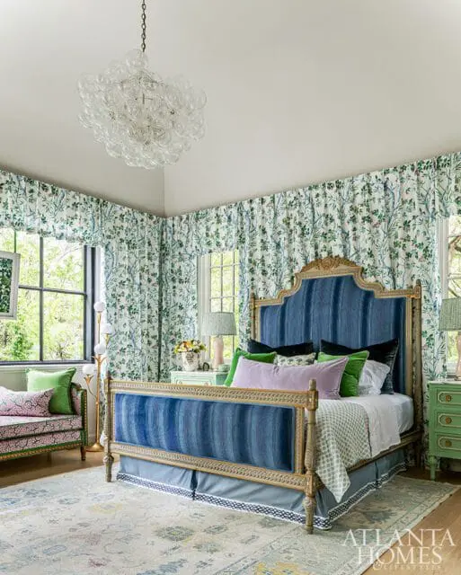 Primary bedroom with a blue upholstered bed, floral curtains, green nightstands, and a patterned rug. A glass chandelier hangs from the ceiling.