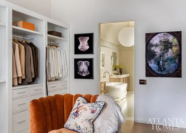 White closet with hanging suits and shirts, an orange velvet chair with decorative pillows, and a view into a bathroom with a freestanding tub.