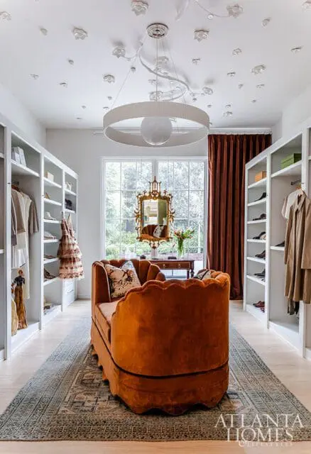 Walk-in closet with white built-in shelving, an orange velvet scalloped sofa, a patterned rug, and a large window with brown curtains.