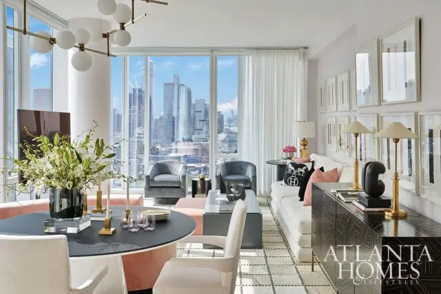 Living room with large windows overlooking a city skyline, featuring a white sofa, velvet armchairs, a round dining table with floral arrangement, and a dark wood credenza with the Atlanta Homes & Lifestyles logo.