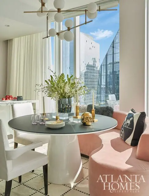 Dining room with a round dark table, white chairs, and a pink curved banquette. A modern chandelier hangs above, and a large window offers city views.