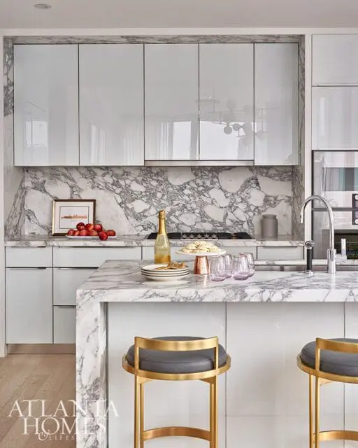 Modern kitchen with white high-gloss cabinets, dramatic marble backsplash and island, and gold-framed bar stools with gray seats.